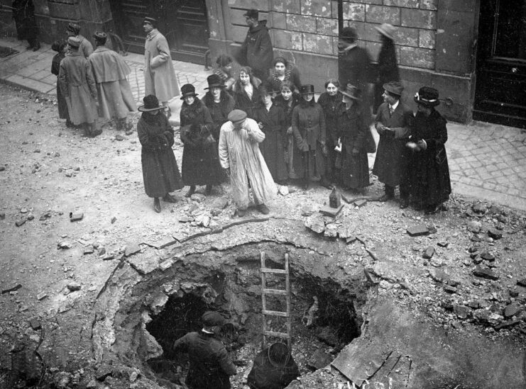 Civilians standing around a bomb crater in the middle of a city street