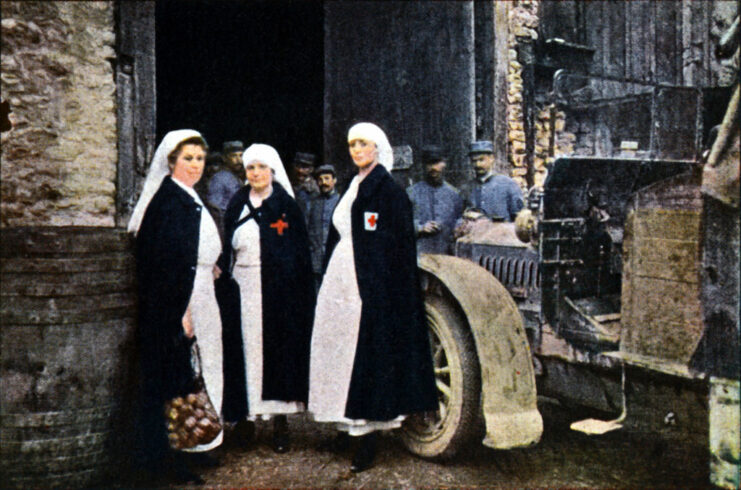 Three Red Cross nurses standing next to a vehicle, with soldiers gathered in the background