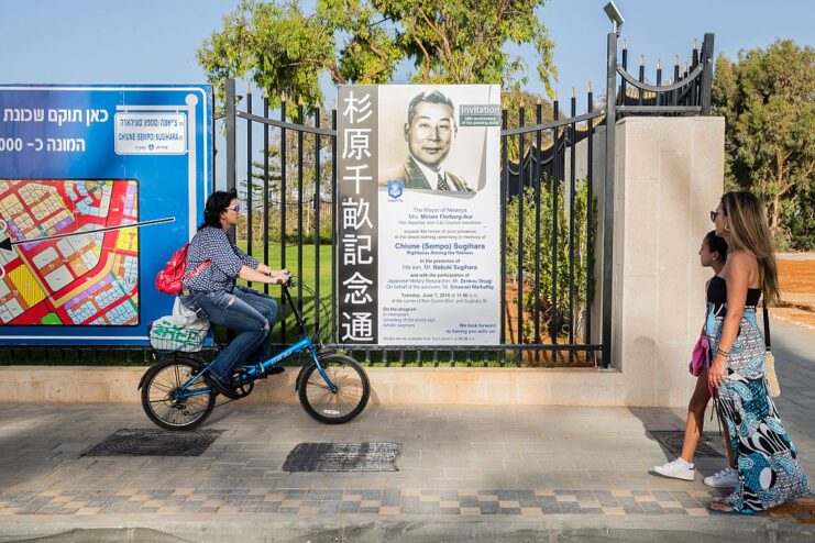 One woman cycling and two women walking past a placard dedicated to Chiune Sugihara