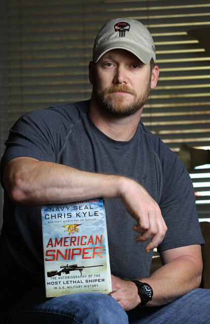 Chris Kyle sitting down, while balancing a copy of 'American Sniper: The Autobiography of the Most Lethal Sniper in U.S. Military History' on his lap