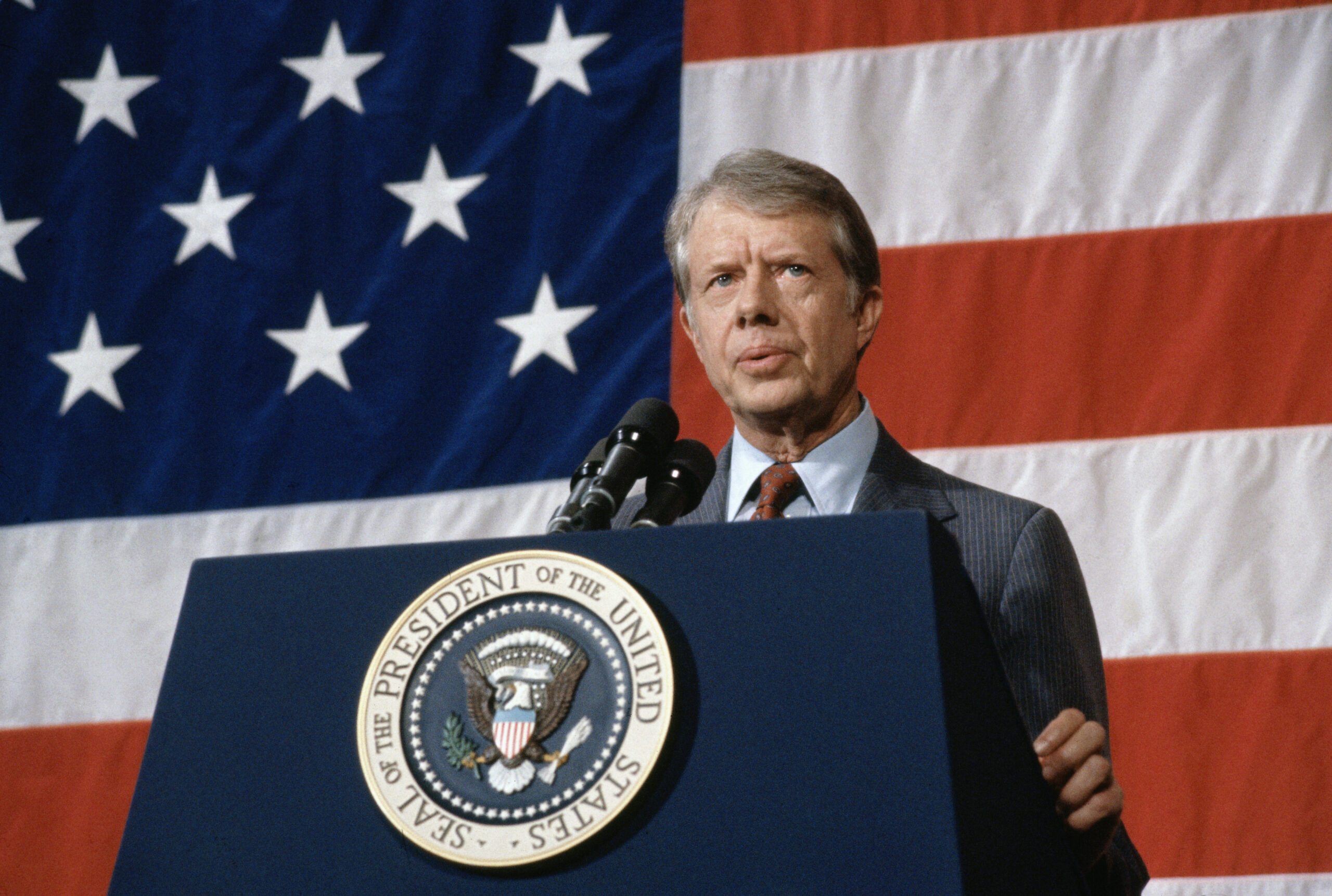 President Jimmy Carter addressing a town meeting. American flag is hung in background.