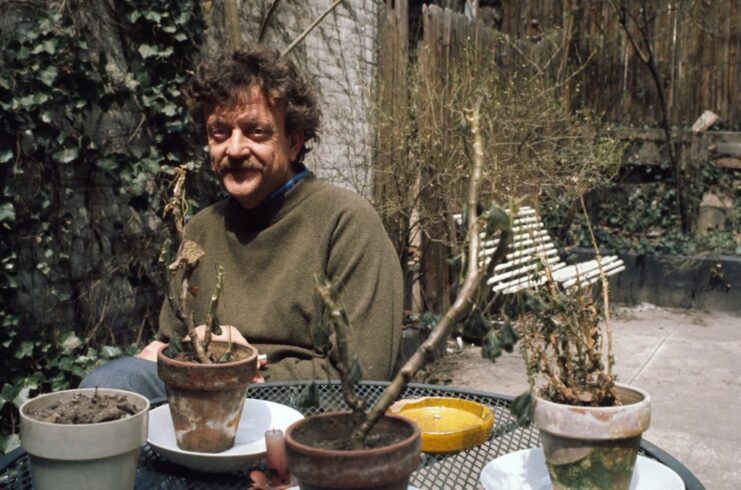 Kurt Vonnegut, Jr. sitting at an outdoor table with potted plants atop it