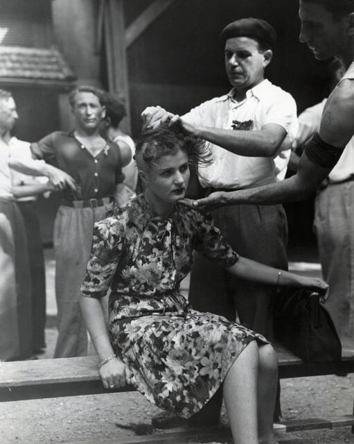 Black and white photo of a woman in a floral dress sitting on a bench while a man shaves her head.