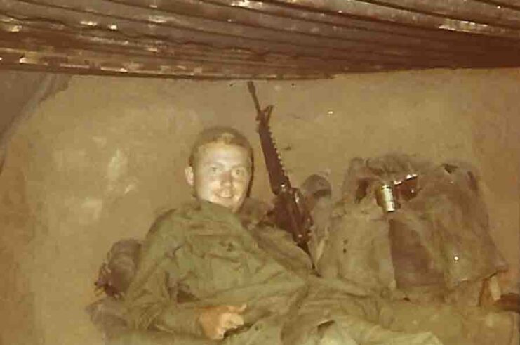 Kenneth J. David leaning on the ground, with his weapon against the wall behind him